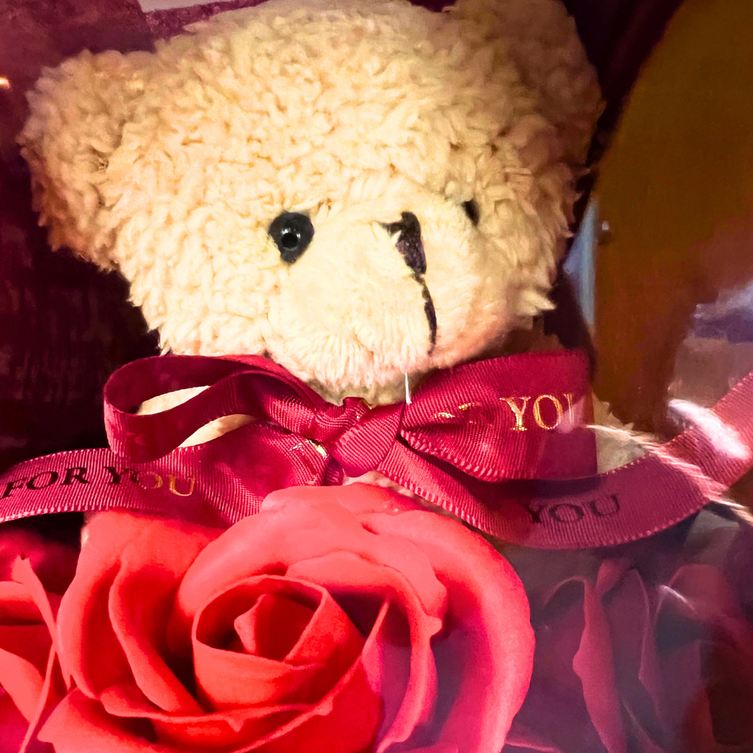 Close-up of a teddy bear with a red ribbon nestled among red soap flowers.