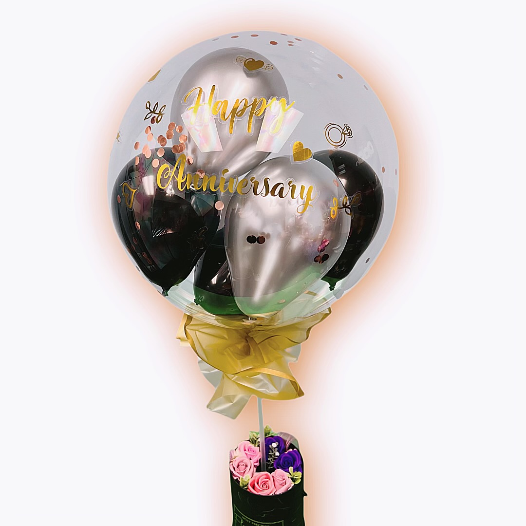 Happy Anniversary BoBo Balloon with gold lettering, filled with black and silver balloons, tied with a yellow ribbon, and paired with a flower arrangement at the base