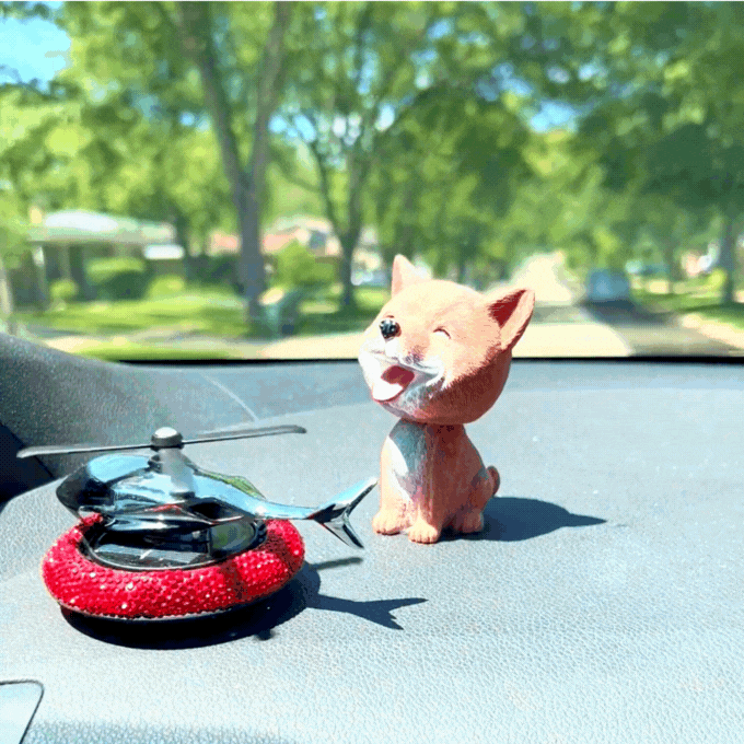 Shaking head dog figurine and a small helicopter decoration on a car dashboard with a sunny outdoor scene in the background.