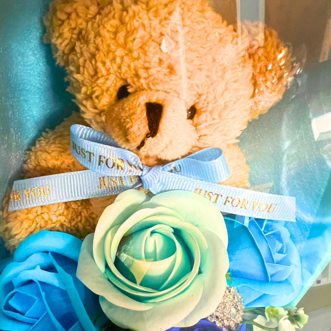 Close-up of a teddy bear with a blue ribbon surrounded by blue and green soap flowers.